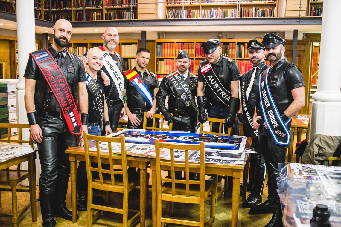 A photo from the opening of the UK Leather & Fetish Archive at Bishopsgate Institute. Eight men are standing round a table in the Library, dressed up in leather.