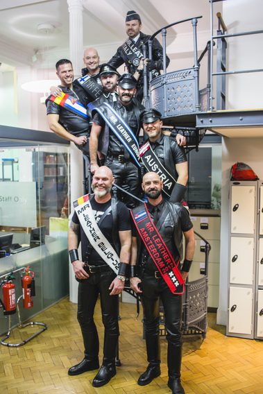 A photo from the opening of the UK Leather & Fetish Archive at Bishopsgate Institute. Eight men are standing up the spiral staircase in the archive.