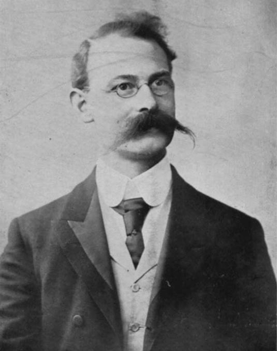 A black and white photo of Charles Goss, who has short hair, round glasses, and a big moustache. He is wearing a suit.