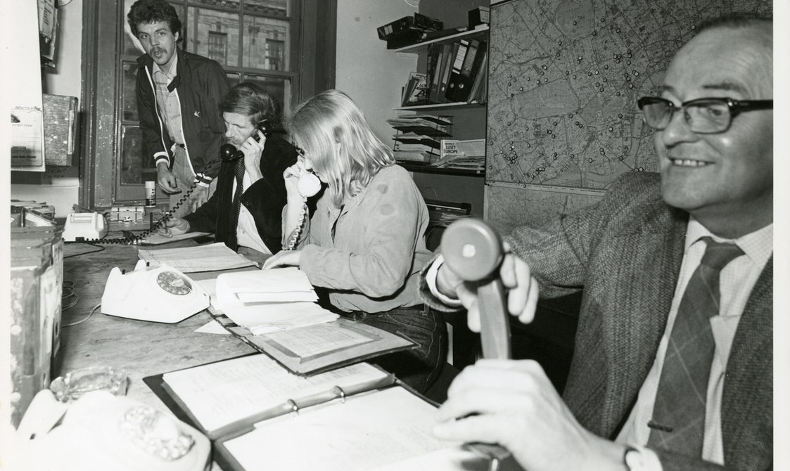 Three people are sitting in a row at the Switchboard office, on the phone to callers. Another person is coming in through the door.