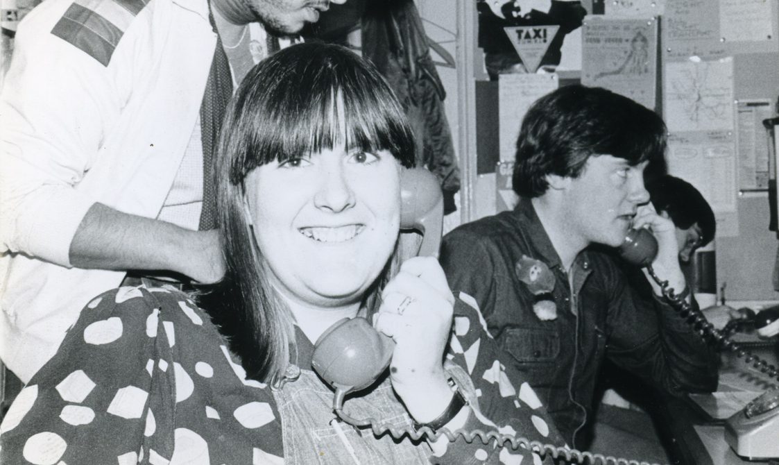 A woman is smiling at the camera, on the phone in the Switchboard office. Other people are on the phone behind her.