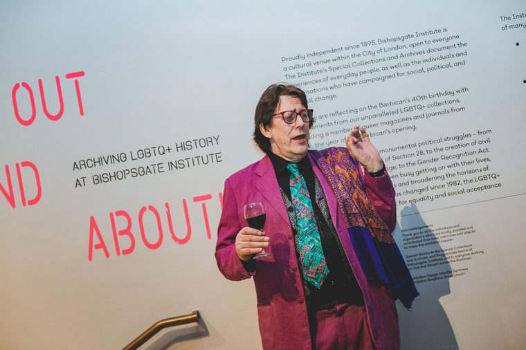 A person in a purple suit and green tie, holding a glass of red wine and looking mock shocked, next to the entrance of the Out and About exhibition.