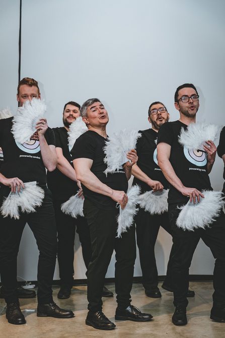 A photograph of members of London's Gay Men's Chorus performing at the exhibition opening. they each are holding a feathered fan in each hand and are using them for emphasis whilst singing.
