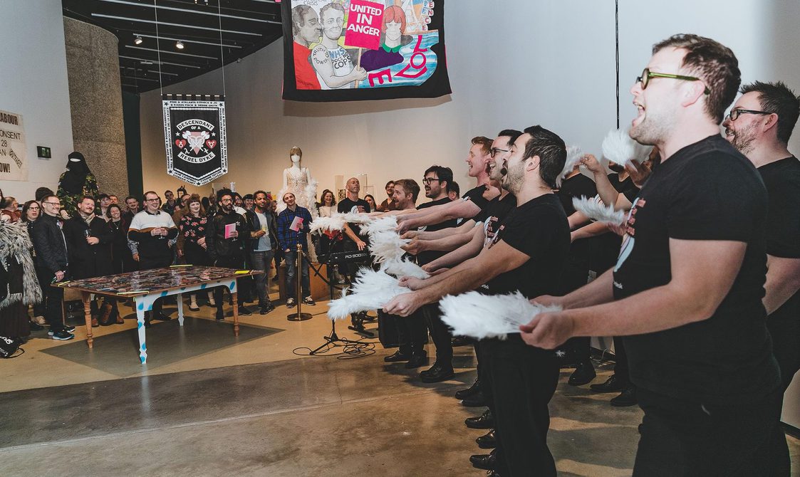 A photograph of members of London's Gay Men's Chorus performing at the exhibition opening. they each are holding a feathered fan in each hand and are using them for emphasis whilst singing in front of a large audience.