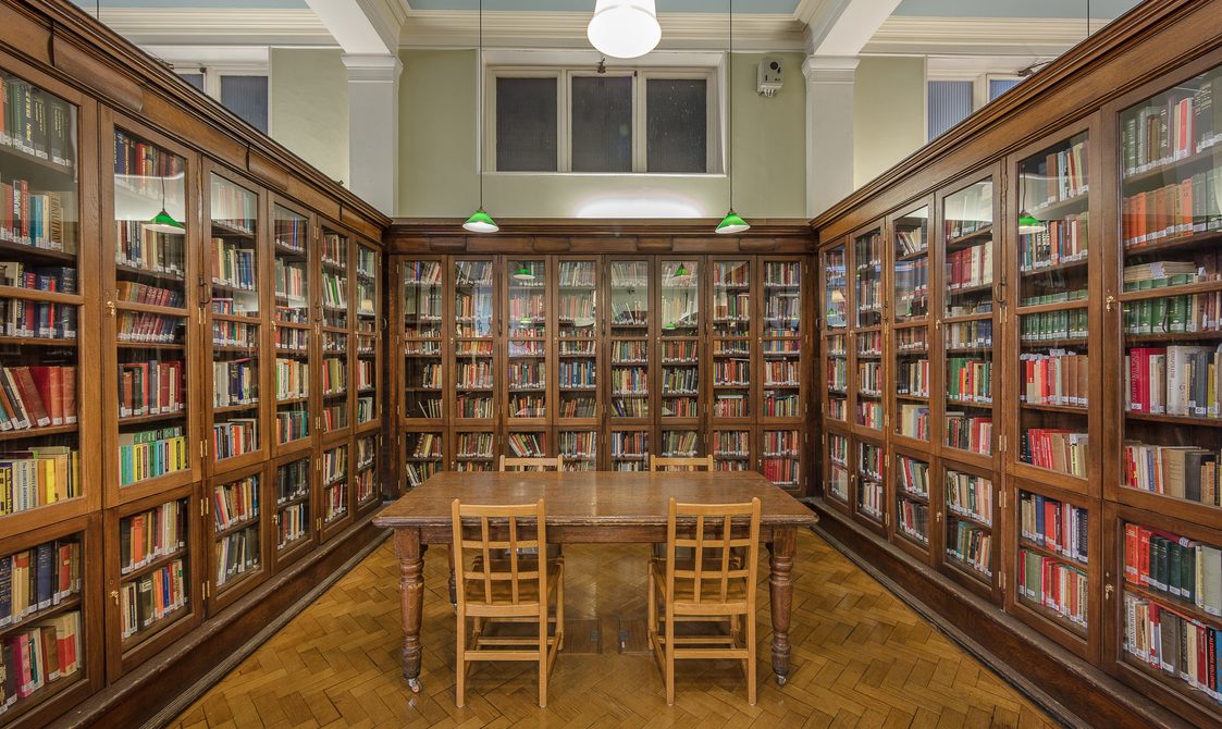 A photo of Bishopsgate Institute Library. Three cabinets with books are in the left hand side, right hand side, and the back of the photo. A wooden desk is in the middle of these cabinets, with four chairs around the table.