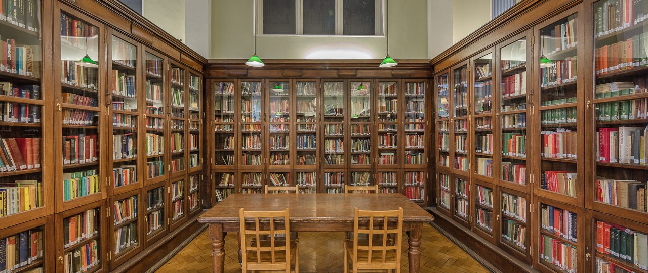 A photo of Bishopsgate Institute Library. Three cabinets with books are in the left hand side, right hand side, and the back of the photo. A wooden desk is in the middle of these cabinets, with four chairs around the table.