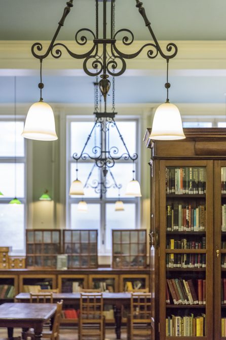 A photo of Bishopsgate Institute library. Books are in wooden cabinets. Lights are dangling from the ceiling, with desks in the centre of the room.