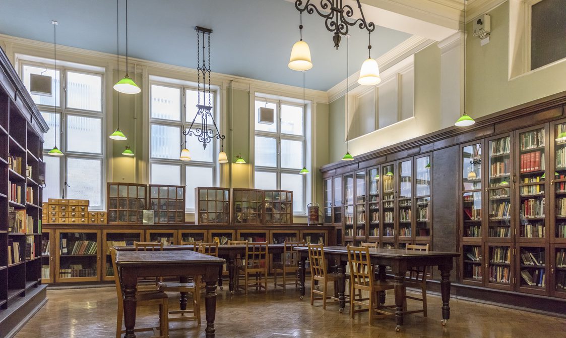 A photo of Bishopsgate Institute Library. Books are in cabinets around the room, with lights hanging from the ceiling. Desks are in U-shape in the centre of the room.