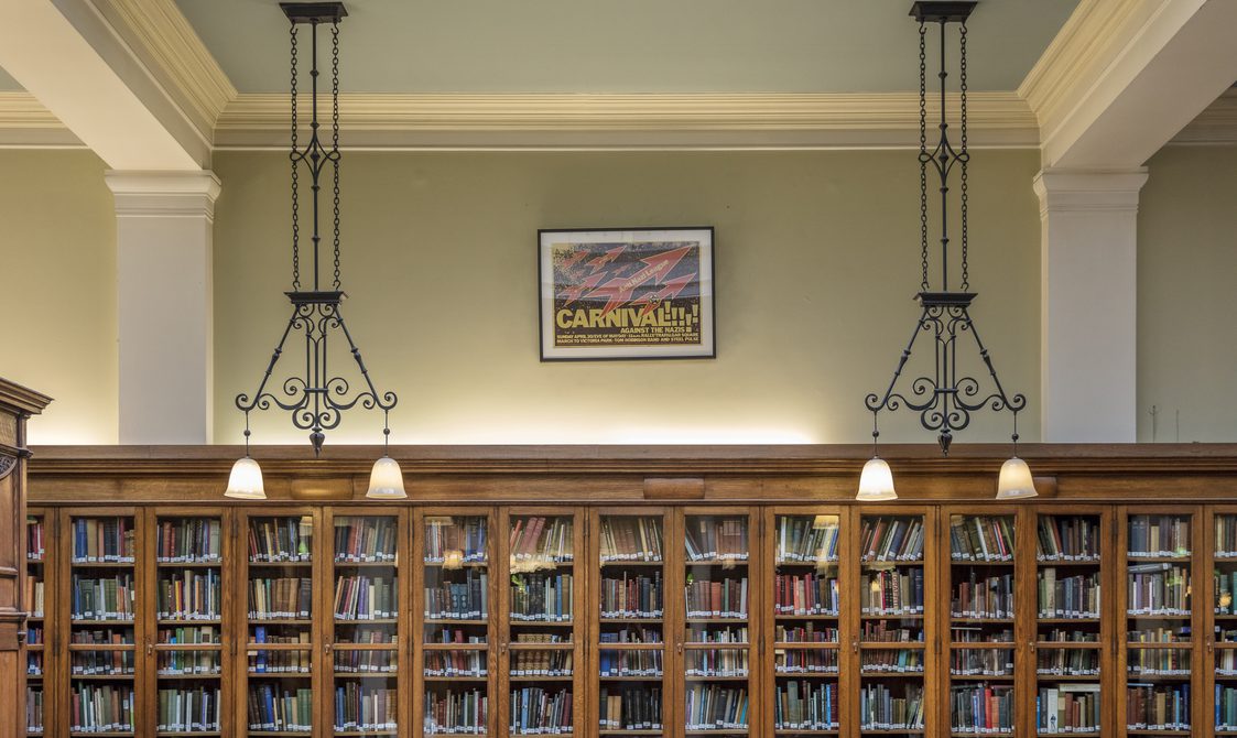 A landscape picture of Bishopsgate Institute Library. Books are in cabinets along the edge of the wall and two lights are hanging from the ceiling.