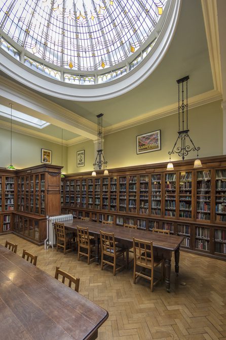 A portrait photo of Bishopsgate Institute library. The ceiling is domed. with books in cabinets along the edge of the room and desks in the centre.