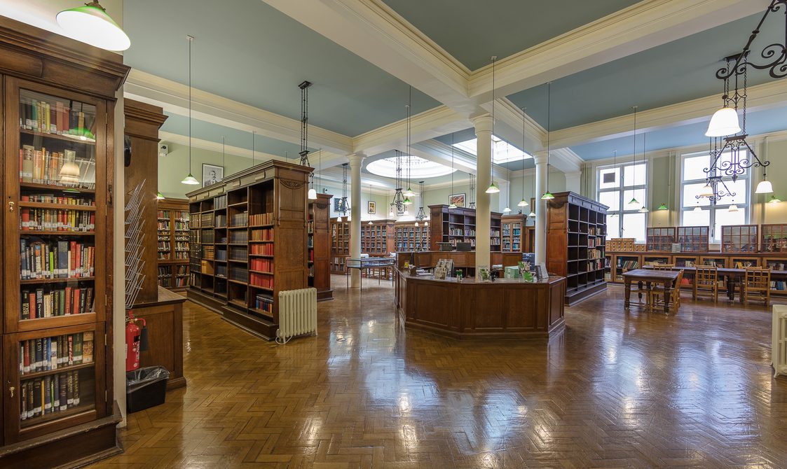 A landscape photo of Bishopsgate Institute Library. The floor is laminate wood flooring, with books in cabinets around the edges of the room. A reception desk is in the centre.