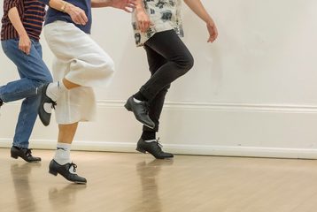 A tap dancing class at Bishopsgate Institute. This photo is of three students who are practising. They are all wearing tap shoes and doing the same move where their left leg is on the ground and they kick out their right leg behind them.