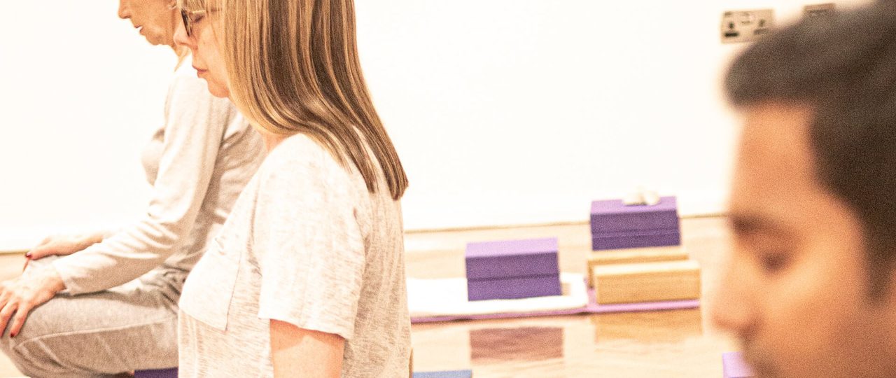 A photo from a Iyengar yoga session at Bishopsgate Institute. The students are sitting crossed legged with their eyes closed and hands on their knees.