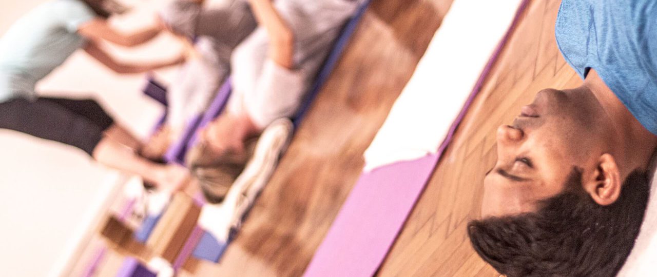 A photo from an Iyengar yoga workshop at Bishopsgate Institute. Students are lying down on their mats, and the teacher is going round, adjusting their posture.