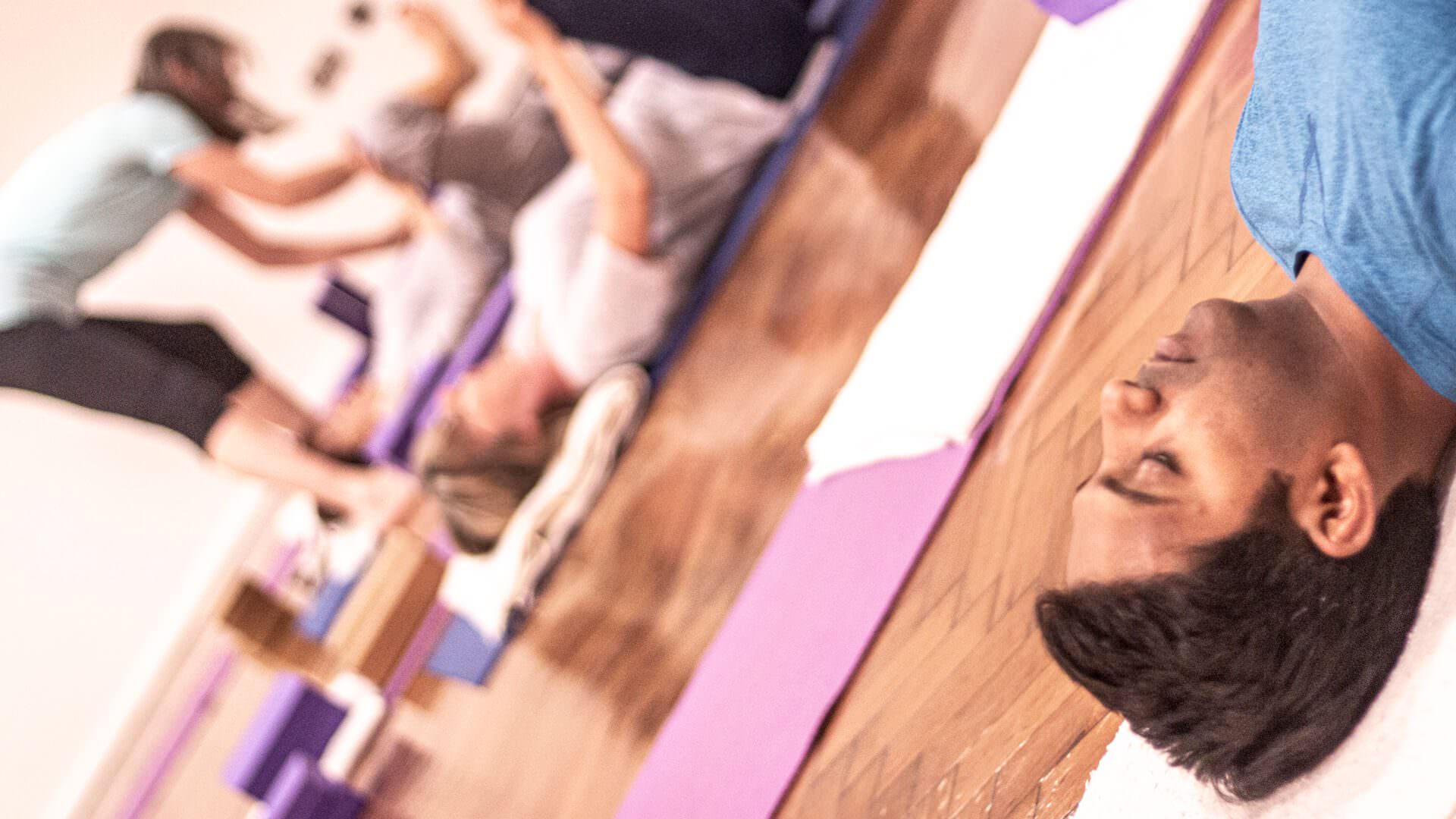 A photo from an Iyengar yoga workshop at Bishopsgate Institute. Students are lying down on their mats, and the teacher is going round, adjusting their posture.