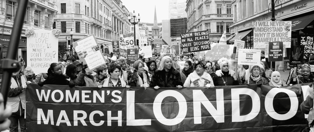 A black and white of the Women's March in London. The women at the front of the march are holding a big banner which reads: "Women's march London".