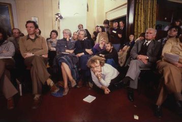 A group of people sitting in rows, with leaflets in their hands. They are looking expectantly to the front of the room. This image is from the History Workshop Events Archive.