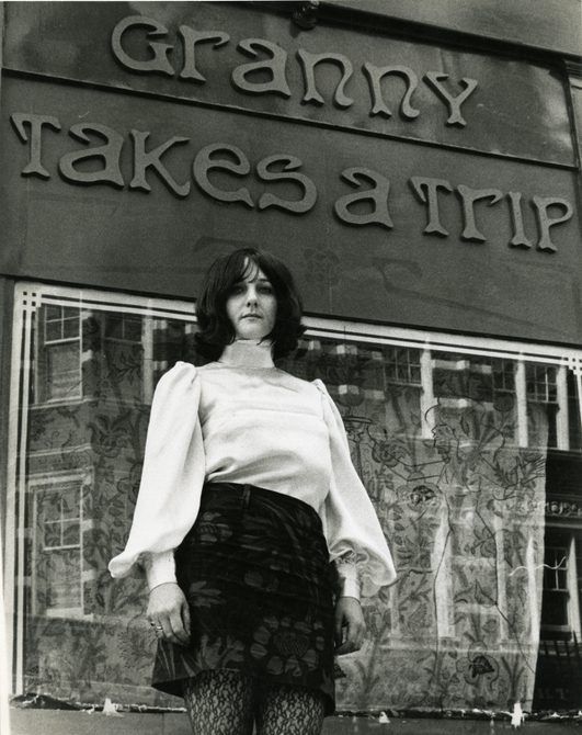 A photo of a woman standing outside the shop Granny Takes a Trip in the 1960s. She is wearing a white turtleneck and black mini skirt, and has dark hair to her shoulders.