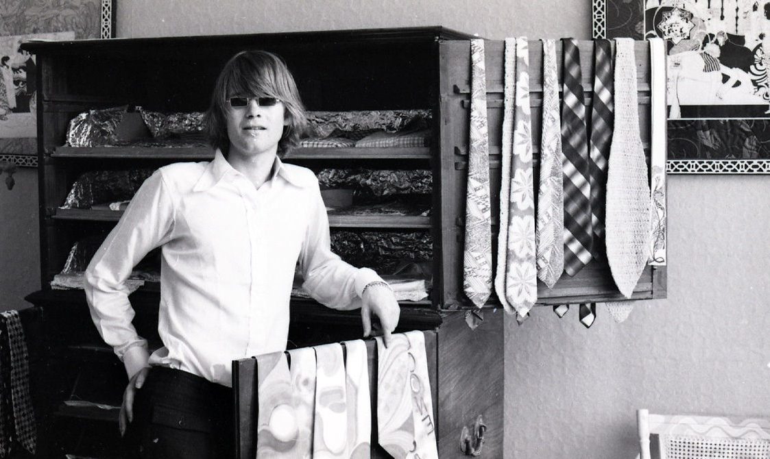 A photo of a man inside the Hung on You shop, he is standing in front of a cabinet of ties. The image is in black and white and taken in the 1960s.