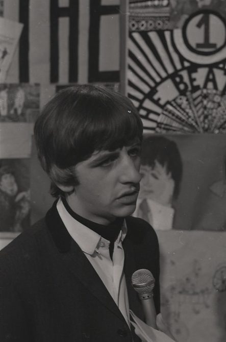 A black and white photo of Ringo Starr being interviewed in the 1960s. A sign behind him says "The Beatles".