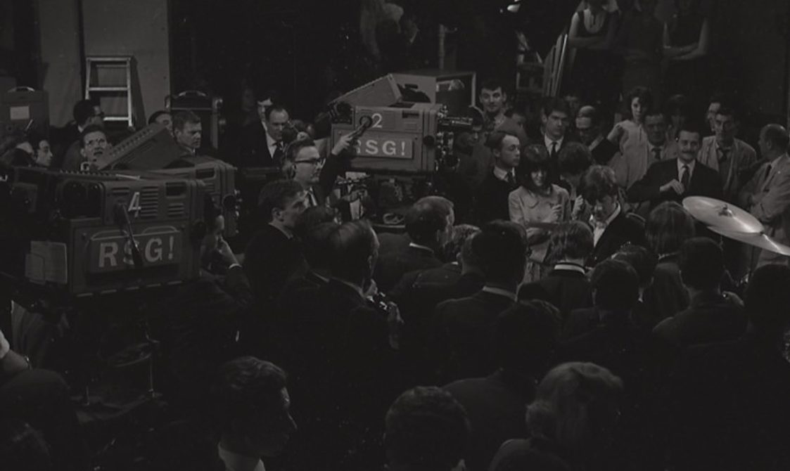 An image of a Beatles concert in the 1960s. The photo is black and white and there are also film camera filming the performance.