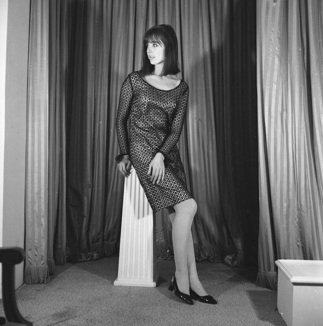 A photo taken at Cash Fashions in the 1960s of a woman in a knee length dress sitting on a plinth. She is turning towards her right, looking away from the camera.