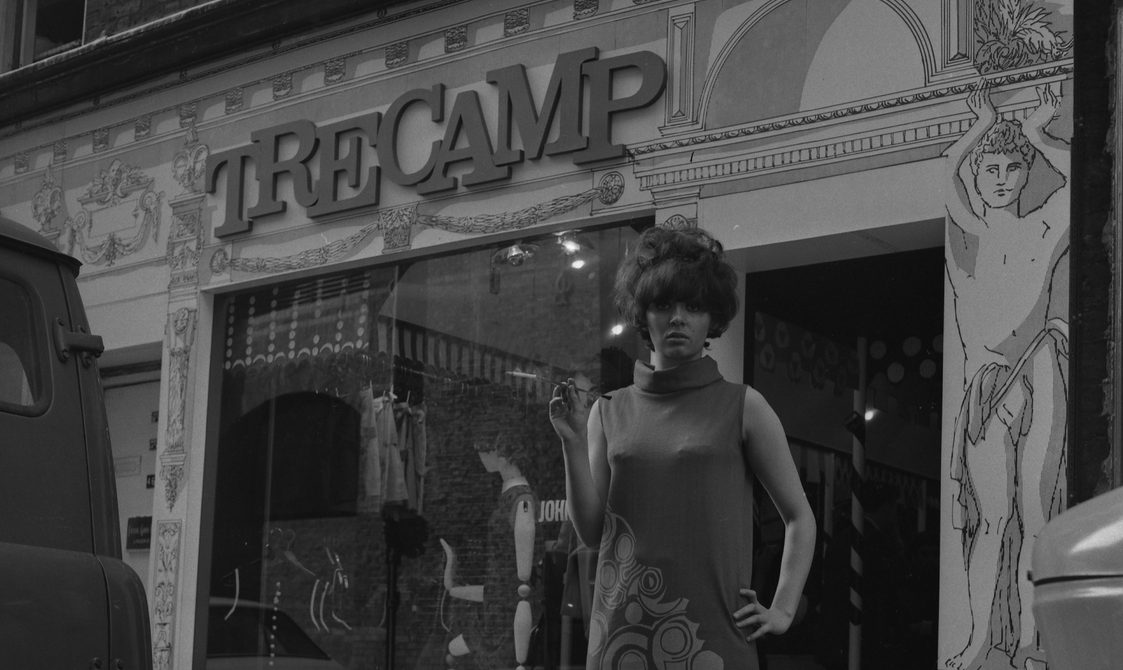 A woman is standing outside the shop Tree Camp in the 1960s. She is wearing a long dress and has her left shoulder on her left hip and is holding a cigarette holder in her right hand.