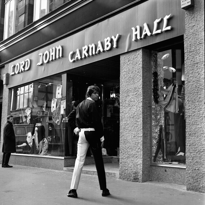An image taken in 1966 on Carnaby Street of a man starting in front of "Lord John Carnaby Hall". He is facing away from the camera, wearing trousers where one leg is white and one black. He has turned his head to look back at the camera.