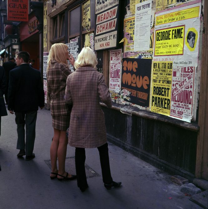 Two woman are looking at posters on Carnaby Street. The posters are mostly advertising concerts. The woman are wearing long coats - one brown and one chequered - and have blonde hair. They are facing away from the camera and towards the posters.