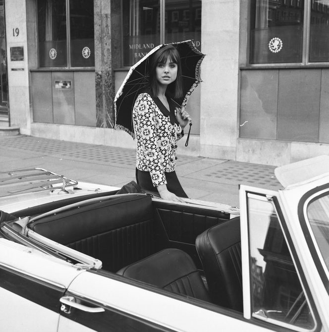 A woman is standing in front of a car, wearing a top with a circular pattern, a black skirt, and is holding an umbrella. The image is in black and white and taken during the 1960s.