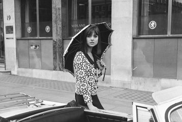 A woman is standing in front of a car, wearing a top with a circular pattern, a black skirt, and is holding an umbrella. The image is in black and white and taken during the 1960s.