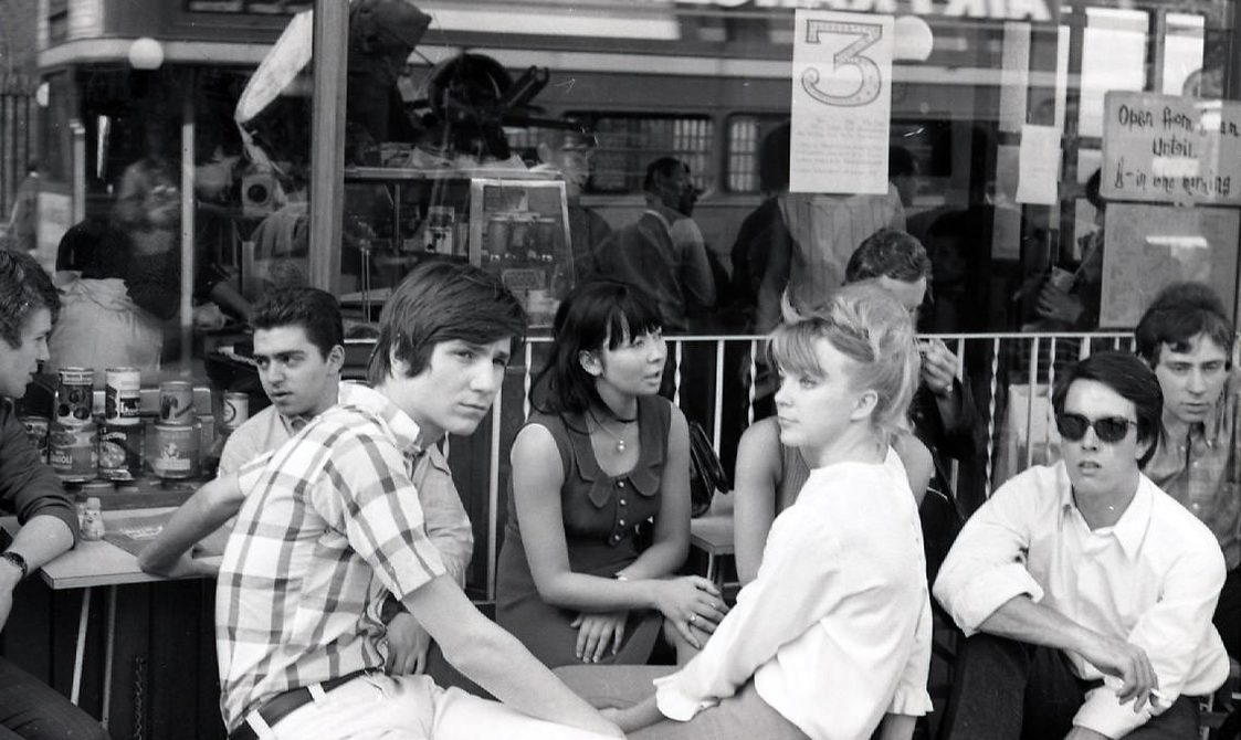 A group of young people are sitting outside a cafe. The image is black and white and was taken in the 1960s by Derek Brook.
