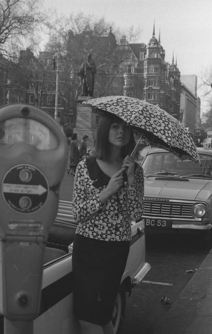 A photo taken outside Cash Fashion sin the 1960s of a woman in a black skirt, floral top, and a matching floral umbrella.
