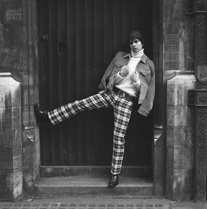 A man is standing in a doorway, wearing chequered trousers, a turtleneck top and a jacket. He has his right leg kicked up in the air. The image is black and white and was taken in the 1960s.