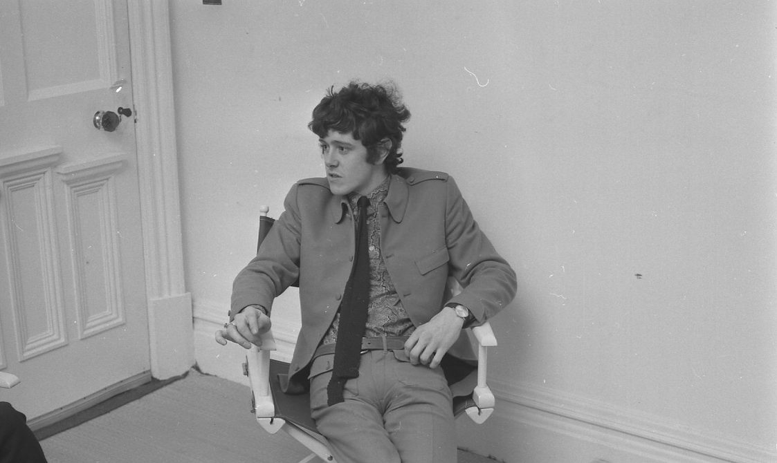 A black and white photo of musician Donovan sitting in a directors chair. He is wearing a suit, tie, and black shoes. He is talking to someone whose legs are shown in the photo.