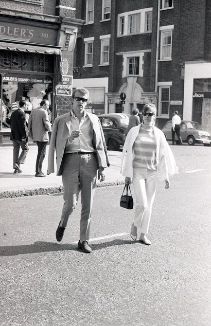 A black and white photo of a man and a woman crossing the road in King's Road in June 1966. They are both wearing trousers and t-shirts, with jackets hung over their shoulders.