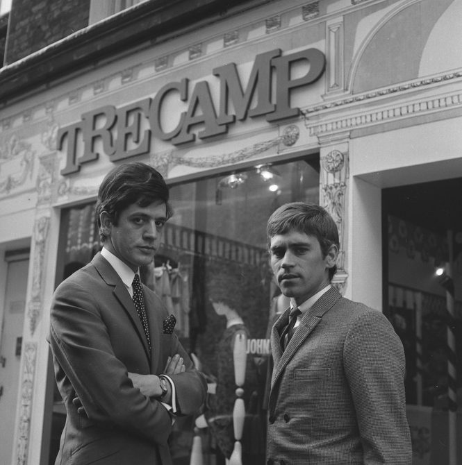 A black and white photo of two men stood outside the shop "Tree Camp" in the 1960s. They are both wearing suits and looking at the camera.