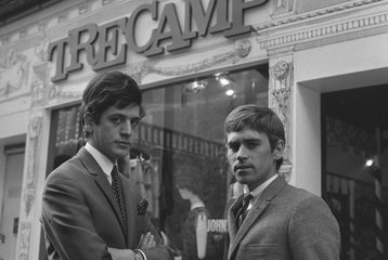 A black and white photo of two men stood outside the shop "Tree Camp" in the 1960s. They are both wearing suits and looking at the camera.