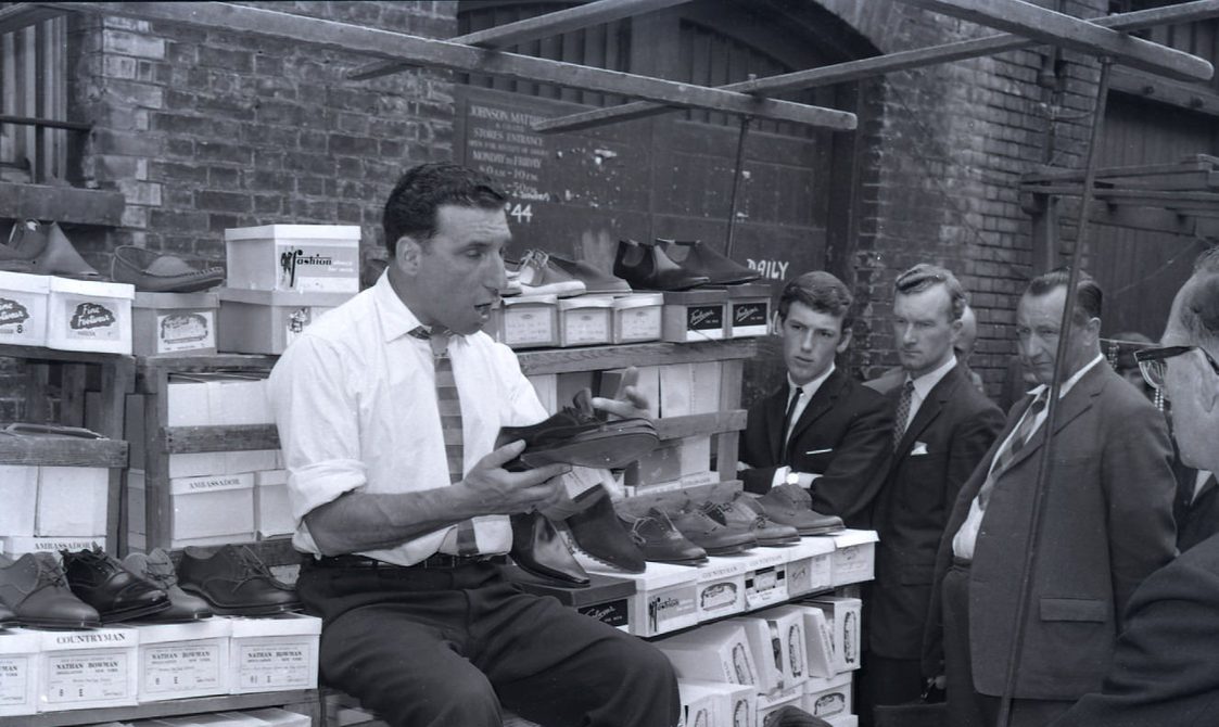 A black and white photo of a man selling shoes in Leather Lane in the early 1960s. He has shoes in his hands and is talking to customers.