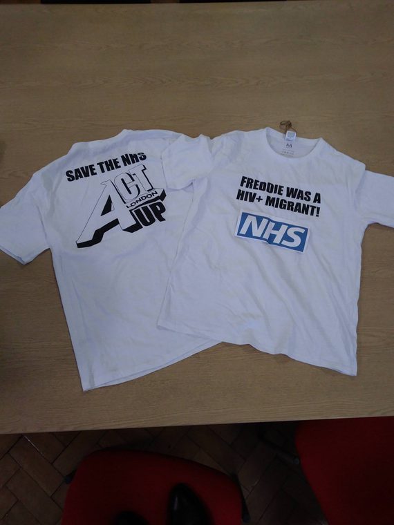 Two white t-shirts are on a desk. The t-shirt on the left says "Save the NHS, ACT UP", and the one on the right says: "Freddie was a HIV+ migrant" and then has the NHS logo underneath.