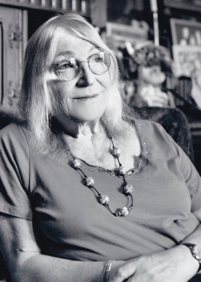A black and white photo of an older woman, sitting down, with shoulder length hair and glasses. She's looking into the distance.