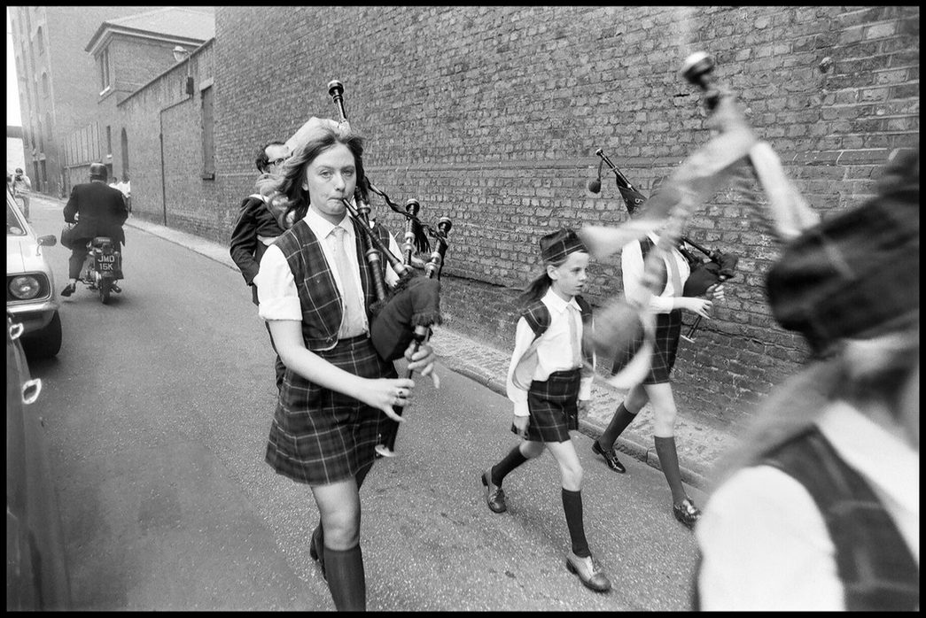 A black and white photograph of a group of girls parade down a small side street, wearing plaid and playing bagpipes.