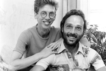 A black and white photo of two men looking at the camera and smiling. One of them is leaning on the other.