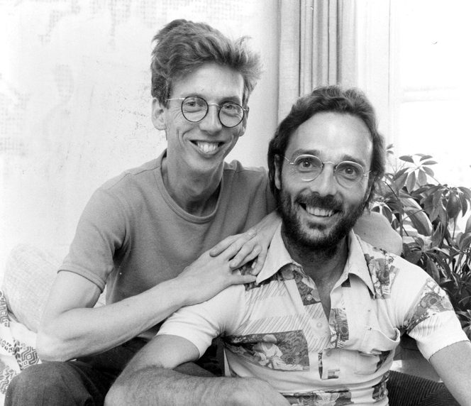 A black and white photo of two men looking at the camera and smiling. One of them is leaning on the other.