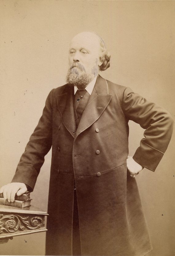 A sepia photo of radical George Howell. He is standing up, wearing a suit and long coat, with his left hand on his hip.