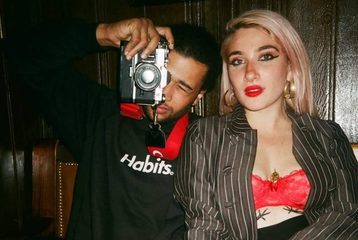 The two people who run London Vagabond. The man on the left is taking a photo, and the woman is sitting next to him, both looking at the camera.