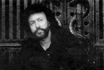 Bearded person with curly hair, wearing dark clothing, leans against an ornate wooden backdrop with hands in pockets.