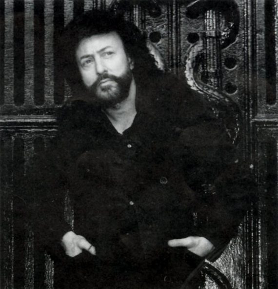 Bearded person with curly hair, wearing dark clothing, leans against an ornate wooden backdrop with hands in pockets.
