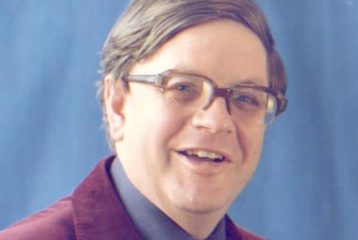 A photo of Norman Longmate, who is wearing a Burgundy blazer and square glasses. He is standing against a blue background.