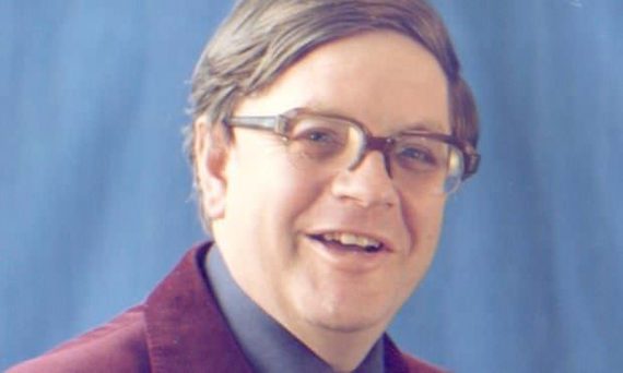 A photo of Norman Longmate, who is wearing a Burgundy blazer and square glasses. He is standing against a blue background.
